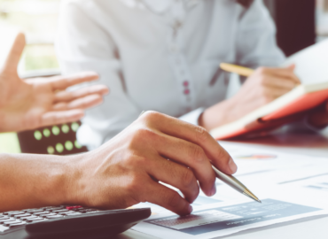 escena de treball en una taula, es veuen les mans amb un bolígraf, una calculadora, i una noia que anota en una llibreta