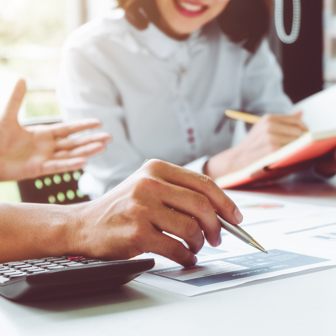 escena de treball en una taula, es veuen les mans amb un bolígraf, una calculadora, i una noia que anota en una llibreta