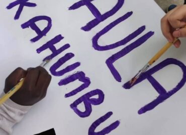 Retall stories. Part superior "Aquest mes de març la temàtica que treballarem a l'espai migdia és Kenya" A baix, "i el personatge és una gran dona: Paula Kahumbu, biòloga i conservacionista de la fauna keniata". La imatge deixa veure com s'està fent una pancarta amb el nom de Paula Kahumbu amb llestres liles sobre blanc. Es veuen les mans de dos infants, que tracen les lletres.